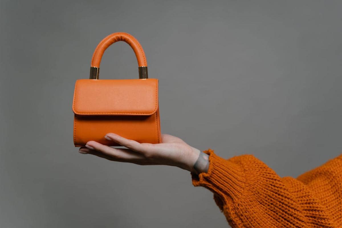 Tangan dengan lengan berbahan rajut memegang tas tangan oranye bergaya dengan pegangan pendek di latar terang.