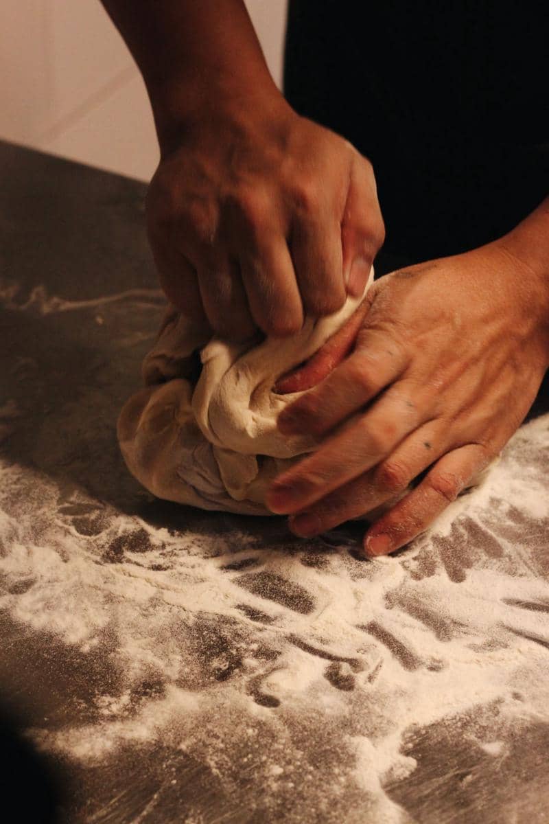Tangan seseorang menguleni adonan roti di atas permukaan dapur sebagai ilustrasi proses membuat roti.