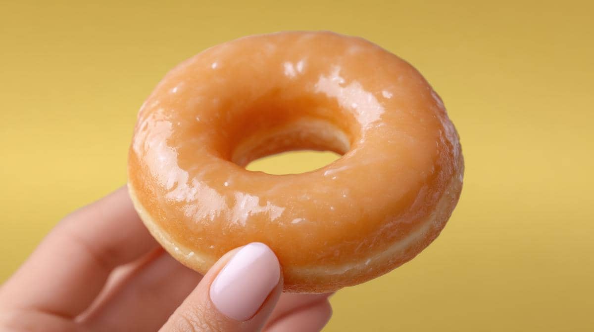 Tangan memegang donat bulat berlapis glasir tanpa topping di depan latar belakang gradasi merah muda dan oranye cerah.